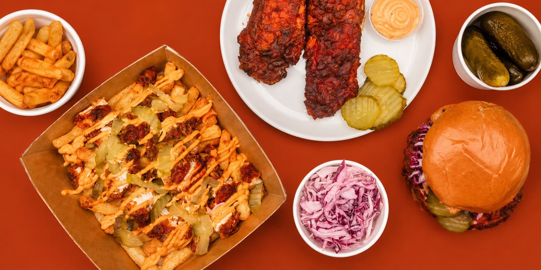 Yaab berbere loaded fries, chicken tenders, burger, slaw and pickles spread on burnt orange background